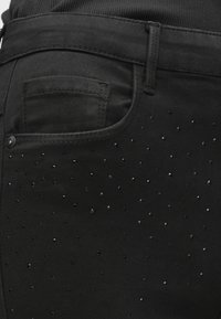 Μαύρο τζιν από ελαστικό ύφασμα, με μια τσέπη και διάσπαρτες μαύρες πούλιες για υφή και λεπτομέρεια κατά μήκος της πλευράς.