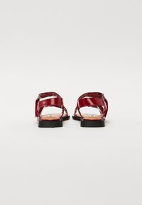 Red patent leather sandals featuring a strappy design, flat sole, and adjustable buckle. Smooth texture with a brown insole.