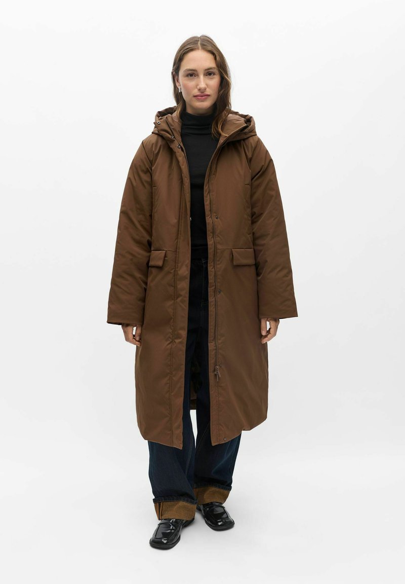 Femme debout portant un long manteau marron à capuche, un col roulé noir, un pantalon large foncé avec des revers et des chaussures noires sur un fond blanc uni.