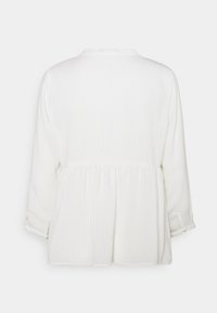 Blusa peplum bianca con maniche lunghe, caratterizzata da una texture a coste e un colletto rotondo; la vista posteriore mostra un dettaglio di vita arricciata.