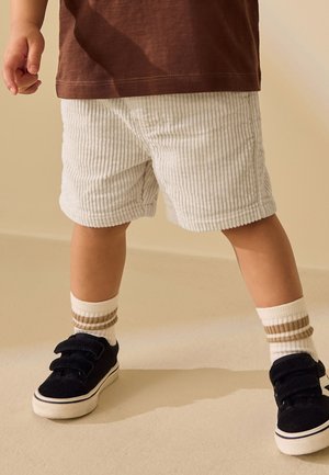 Kleinkind trägt weiße gerippte Shorts, braunes Shirt, gestreifte weiße Socken und schwarze Klett-Sneakers und steht auf einem beigen Boden.