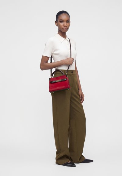 Femme portant un top blanc à manches courtes et un pantalon large couleur olive, tenant un petit sac à main rouge avec une bandoulière noire sur l’épaule.