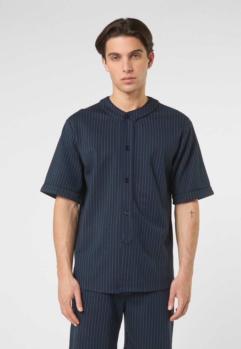 Junger Mann mit kurzen dunklen Haaren, der ein dunkelblaues, kurzärmeliges Hemd mit Nadelstreifen und passende Hose trägt, steht neutral da.