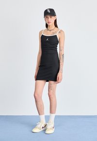 Zwarte aangesloten jurk met witte bandjes en een klein Jordan-logo. Draagt een zwarte pet en beige sneakers met rode accenten.