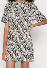 Schwarzes und weißes Kurzarmkleid mit floralem Muster und vertikalen Stängeln, hohem Ausschnitt und lockerer Passform. Weiche Stofftextur.