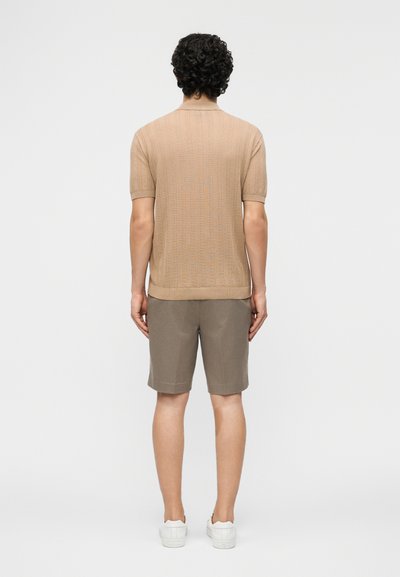 Mann mit lockigem Haar, der ein beiges kurzärmeliges Strickpoloshirt, taupefarbene knielange Shorts und weiße Turnschuhe trägt, steht mit dem Rücken zur Kamera.