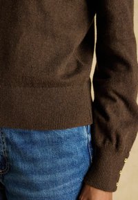 Brauner gestrickter Pullover mit geripptem Saum und Bündchen, verziert mit goldenen Knopfakzenten an den Ärmeln, kombiniert mit blauen Jeans.