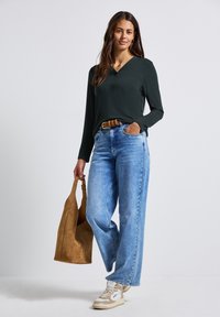 Haut à manches longues vert foncé avec un col en V, jeans taille large bleu clair, et un sac à main en daim marron. Des baskets blanches et beiges complètent la tenue.