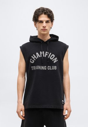 Young man with curly hair wearing a sleeveless black hoodie with "CHAMPION TRAINING CLUB" text, standing against a plain light background.