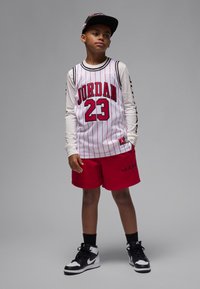 Maillot de basketball blanc avec des rayures rouges et inscriptions « JORDAN 23 », accompagné d'un short rouge et de baskets noires, avec une casquette portée sur la tête.