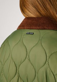 Zelená prošívaná bunda s hnědým kordovým límcem a černou nášivkou "Pepe Jeans" na zadní straně. Na látce je viditelný texturovaný vzor.