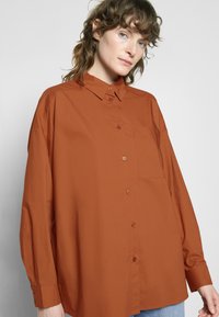 Camisa castanha de botões, feita de tecido suave, com colarinho, mangas longas e um bolso no peito, exibida contra um fundo neutro.