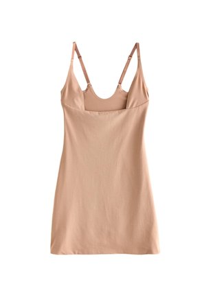 Jonge vrouw met licht haar in een beige nauwsluitende mouwloze jurk, staand met één hand op haar heup tegen een effen lichte muur.