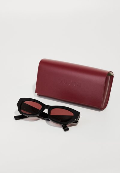 Gafas de sol negras con montura angular y lentes tintados en rojo, acompañadas de un estuche de cuero burdeos embellecido con "GUCCI".