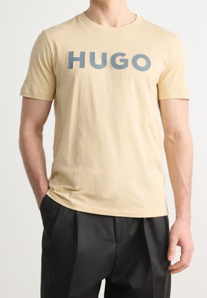 Beżowy t-shirt z krótkim rękawem z ciemnoniebieskim napisem "HUGO", noszony z czarnymi plisowanymi spodniami, ręce luźno po bokach.