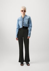 Cropped denim jacket in light blue with button closure and two chest pockets, paired with high-waisted black flared pants and black slide sandals.