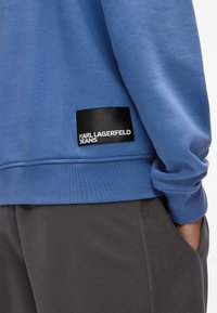 Modrá mikina se žebrovanými manžetami a lemem, která má na levé straně černou cedulku s bílým textem "KARL LAGERFELD JEANS".