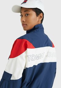 Marineblå, rød og hvid farveblokeret jakke med "TOMMY JEANS" logo på ryggen. Har en høj krave og sidelommer. Glat, letvægtsstof.
