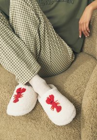 Weiße, flauschige Hausschuhe mit rotem Hummer-Stickerei; kombiniert mit hellgrauen Socken und grün-karierten Hosen, auf einer strukturierten beige Oberfläche.