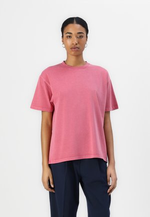 Lyserød oversized t-shirt med kortærmer, rund halsudskæring, blød bomuldsstruktur og subtilt falmet udseende, parret med mørke bukser.