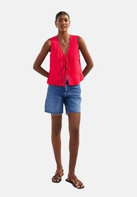 Haut sans manches texturé rouge avec un col en V et une fermeture à nouer, associé à un short en jean bleu clair. Le modèle porte des sandales brunes.