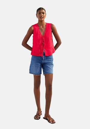 Haut sans manches texturé rouge avec un col en V et une fermeture à nouer, associé à un short en jean bleu clair. Le modèle porte des sandales brunes.