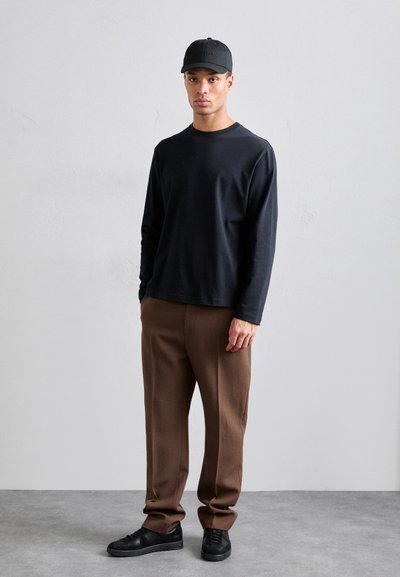 Sort langærmet skjorte, brune smalle bukser, sorte sneakers og en sort kasket. Outiten har en afslappet pasform og et minimalt design.