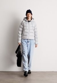 Svijetlosiva jakna s kapuljačom i vezicama, uparena s opuštenim plavim trapericama i crnim klomama, drži crnu torbu.