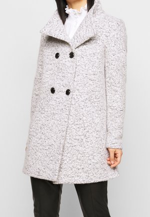 Vrouw draagt een lichtgrijze, getextureerde dubbelgeknoopte jas met zwarte knopen over een zwarte broek en een witte blouse met ruches.