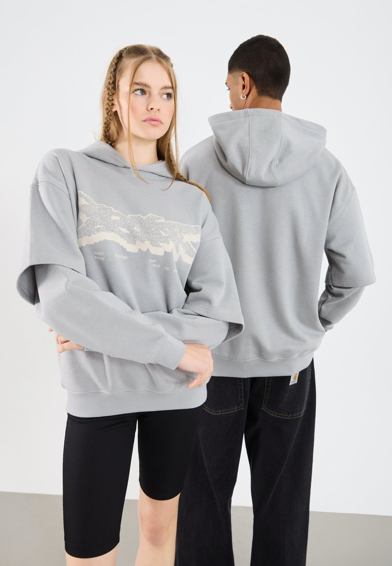 Hoodie gris de gran tamaño con un diseño gráfico en el frontal, acompañado de pantalones cortos negros, en dos modelos que muestran la vista posterior.