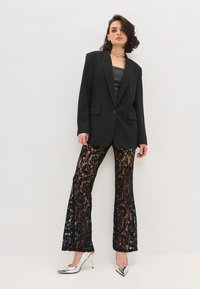 Blazer nero su misura sopra un top in satin nero, abbinato a pantaloni in pizzo nero a campana. Presenta tacchi a stiletto argentati e collane metalliche a strati.