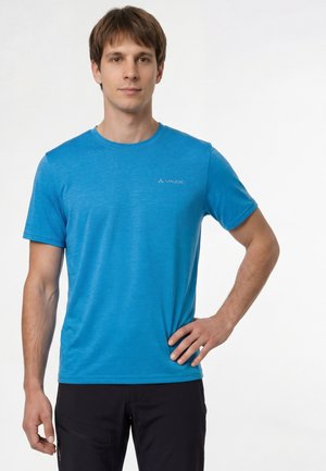 Giovane uomo in piedi con la mano sul fianco, indossa una maglietta blu Vaude e pantaloni neri su uno sfondo bianco semplice.