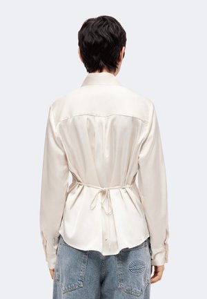 Personne aux cheveux courts et foncés portant un chemisier en satin crème noué à la taille et un jean en denim bleu clair vu de dos.