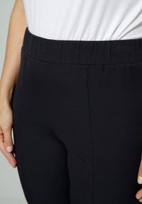 Svarta tygbyxor med elastisk midja, med en slitstark yta och utan synliga knäppningar eller fickor. Närbild på midjan och sidan.