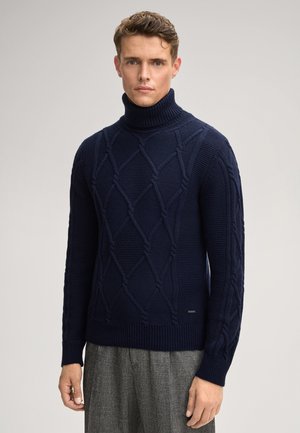 Mann in einem navyblauen, grob gestrickten Rollkragensweater und grauen Wollhosen, der vor einem schlichten hellen Hintergrund steht.