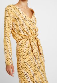 Kvinna iklädd en långärmad klänning i gult och brunt med prickar, knappar och detaljer i knuten midja mot en enfärgad bakgrund.