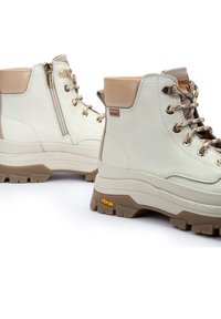 Botas de senderismo de cuero blanco con acentos beige, cordones en la parte delantera, cremallera lateral y suela robusta de Vibram en marrón. Acabado texturizado y diseño resistente.
