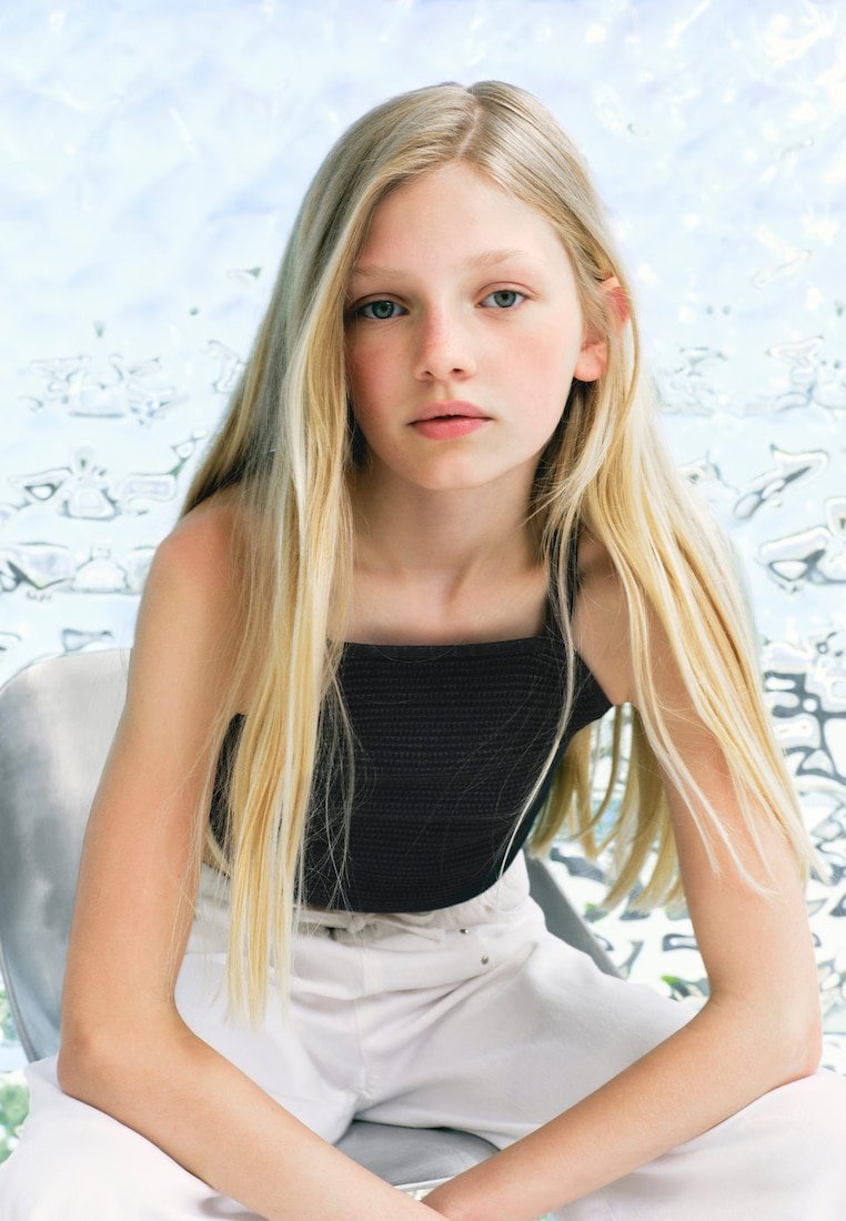 Jeune fille blonde aux longs cheveux, portant un top noir sans manches et un pantalon blanc, assise sur une chaise grise sur un fond flou.
