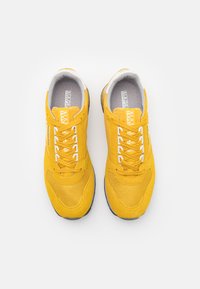 Scarpe da ginnastica gialle realizzate in materiali scamosciati e in rete, con lacci bianchi, una striscia di accento e una suola imbottita. Vista dall'alto.