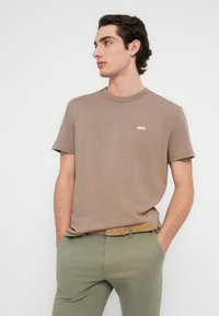 Καφέ κοντομάνικη μπλούζα με στρογγυλή λαιμόκοψη. Το ύφασμα είναι λείο· διαθέτει ένα μικρό κεντητό σχέδιο στο στήθος.