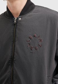 Primer plano de una chaqueta gris oscuro con cremallera, con cuello acanalado y un logotipo circular de texto "SAINT SAL" en el lado izquierdo del pecho.