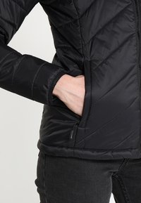 Doudoune noire à fermeture éclair, poches latérales et motifs de couture diagonale. Confectionnée dans un matériau léger et brillant.