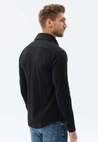 Camicia nera a maniche lunghe con colletto a punta, realizzata in tessuto di trama. Presenta polsini con bottoni e una vestibilità su misura. La vista posteriore mostra i dettagli del design.