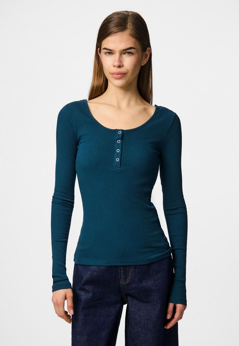 Teal ribbad långärmad topp, har en rundad halsringning och en fyra-knapps placket framtill. Mjuk tyg med åtsittande silhuett, kombinerad med mörka jeans.