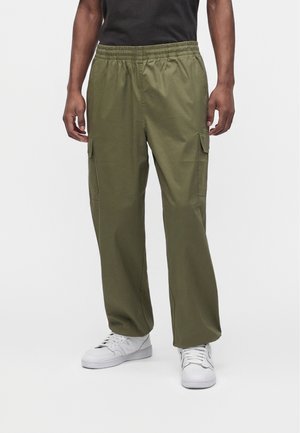 Person trägt olivgrüne Cargohosen mit elastischem Bund und weißen Sneakers, steht vor einem schlichten hellen Hintergrund.