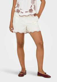 Shorts en lin blanc avec un ourlet festonné et des détails de couture rouge contrastants, associés à des chaussures plates bordeaux et un haut ample.