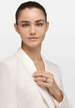 Silver ring with three bands, featuring black and clear accents, worn on a hand against a white blazer with a shawl collar.