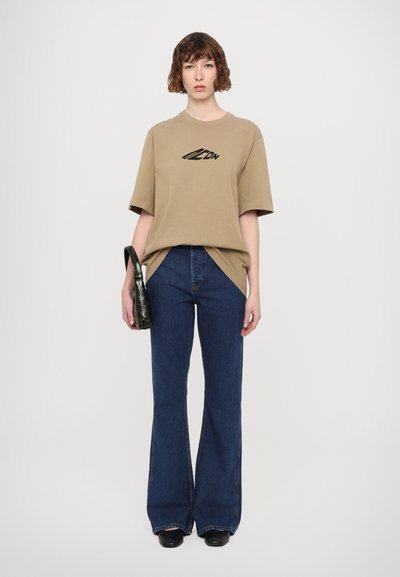 Beige oversized t-shirt met een zwart logo, gecombineerd met blauwe flared jeans en een gestructureerde zwarte handtas. Zwarte schoenen maken de outfit compleet.
