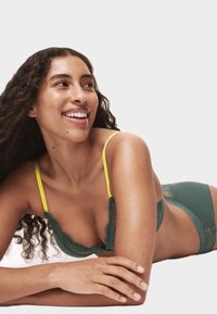 Turkusowy koronkowy biustonosz z żółtymi ramiączkami, z falbankowymi krawędziami i dopasowanymi turkusowymi koronkowymi majtkami. Ciepły odcień skóry i luźne kręcone włosy.