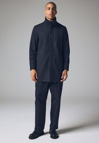 Zwarte wollen jas met een hoge kraag, lange mouwen en twee voorzakken, gecombineerd met een donkere broek en zwarte veterschoenen.